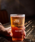A hand holds a clear pint glass with a frosted design featuring a rooster and the text 'Cornish O'cock Craft Cider' against a dark, rustic wooden table background.