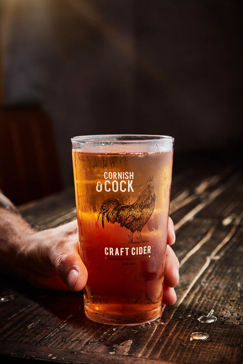 A hand holds a clear pint glass with a frosted design featuring a rooster and the text 'Cornish O'cock Craft Cider' against a dark, rustic wooden table background.