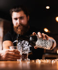 A man with a beard pours a dark amber liquid from a bottle labeled 'Crazy Dog' into two glasses on a wooden bar counter, with a blurred bar interior in the background.