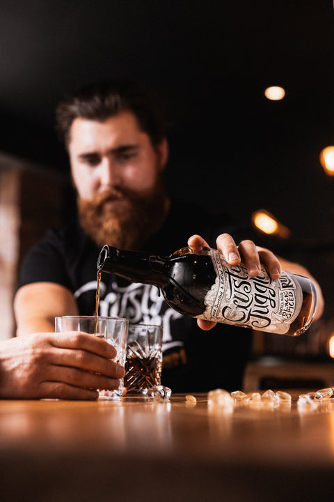 A man with a beard pours a dark amber liquid from a bottle labeled 'Crazy Dog' into two glasses on a wooden bar counter, with a blurred bar interior in the background.