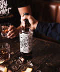 A bottle of Rusty Juggler Straight Bourbon Pineapple Rum stands on a wooden tray beside a glass being filled with amber liquor, set on a rustic table in a dimly lit bar environment.