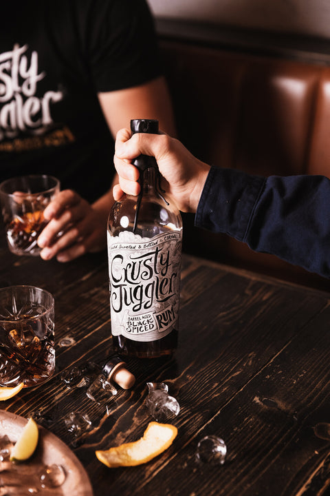 A bottle of Rusty Juggler Straight Bourbon Pineapple Rum stands on a wooden tray beside a glass being filled with amber liquor, set on a rustic table in a dimly lit bar environment.
