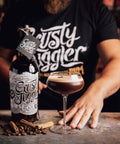 A man in a black graphic t-shirt stands behind a bar counter with a bottle of Rusty Jiggler Spiced Wine and a coupe glass filled with a dark cocktail and foam, with cinnamon sticks on the counter.