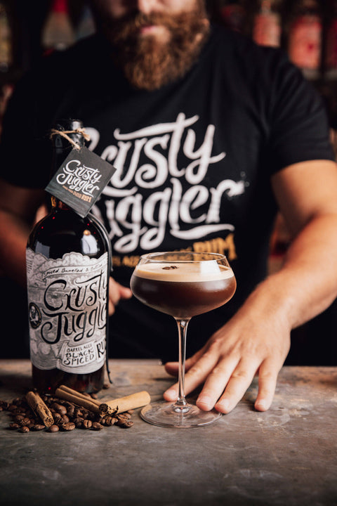 A man in a black graphic t-shirt stands behind a bar counter with a bottle of Rusty Jiggler Spiced Wine and a coupe glass filled with a dark cocktail and foam, with cinnamon sticks on the counter.
