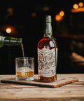 A bottle of Rusty Juggler Straight Bourbon Pineapple Rum stands on a wooden tray beside a glass being filled with amber liquor, set on a rustic table in a dimly lit bar environment.