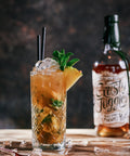 A highball glass filled with an amber cocktail, garnished with mint, a pineapple wedge, and ice, set on a wooden table beside a bottle labeled Rusty Juggler.