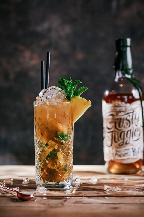 A highball glass filled with an amber cocktail, garnished with mint, a pineapple wedge, and ice, set on a wooden table beside a bottle labeled Rusty Juggler.