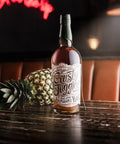 A bottle of Rusty Juggler Buckeye Pineapple Rum sits on a dark wooden bar table, styled with a whole pineapple in the background against a warm, dimly lit bar interior.