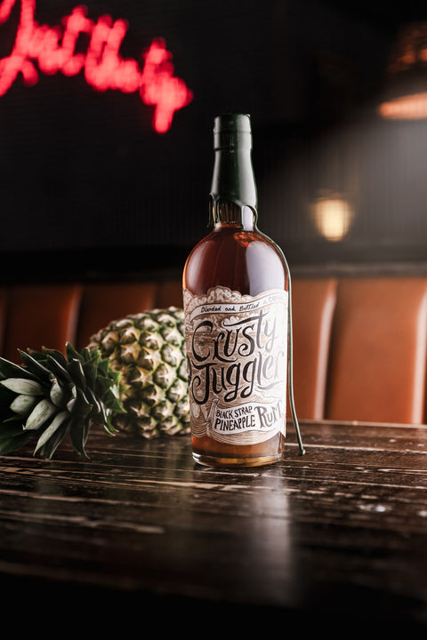 A bottle of Rusty Juggler Buckeye Pineapple Rum sits on a dark wooden bar table, styled with a whole pineapple in the background against a warm, dimly lit bar interior.