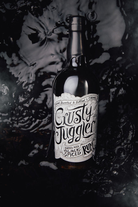 A dark glass bottle of Crusty Juggler Black Spiced Rum with a white label, set against a dark, glossy, water-rippled background.