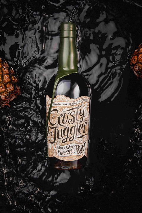 A bottle of Rusty Juggler Black Star Pineapple Rum centered against a glossy black liquid splash background with pineapple crown props on the sides.