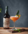 A bottle of Crushed Pineapple Rum with a prepared orange cocktail in a coupe glass, garnished with a green chili and pineapple slice, set on a wooden board against a dark gray background.