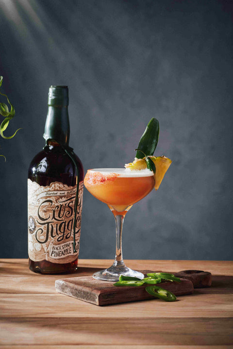 A bottle of Crushed Pineapple Rum with a prepared orange cocktail in a coupe glass, garnished with a green chili and pineapple slice, set on a wooden board against a dark gray background.