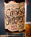 A bottle of Crusty Juggler Hot Honey Rum Liqueur with a rustic paper label, shown on a wooden surface next to a glass of the amber-colored liqueur against a dark background.