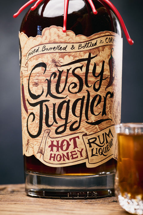 A bottle of Crusty Juggler Hot Honey Rum Liqueur with a rustic paper label, shown on a wooden surface next to a glass of the amber-colored liqueur against a dark background.