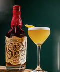 A bottle of Crusty Juggler Hot Honey Rum Cocktail mix stands next to a coupe glass filled with a golden yellow cocktail and a lemon twist, set on a wooden table against a dark green background.