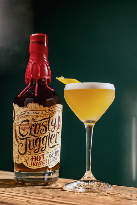 A bottle of Crusty Juggler Hot Honey Rum Cocktail mix stands next to a coupe glass filled with a golden yellow cocktail and a lemon twist, set on a wooden table against a dark green background.
