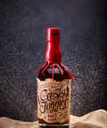 A bottle of 'Crusty Juggler' hot funnel run honey liqueur stands on a textured beige fabric surface against a dark, smoky background with sand-like particles floating around it. The bottle has a red wax seal and a vintage-style label.