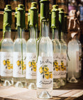 A row of clear glass bottles with white labels featuring yellow lemon illustrations and green stoppers, arranged on a rustic wooden surface with a warm, dimly lit background.