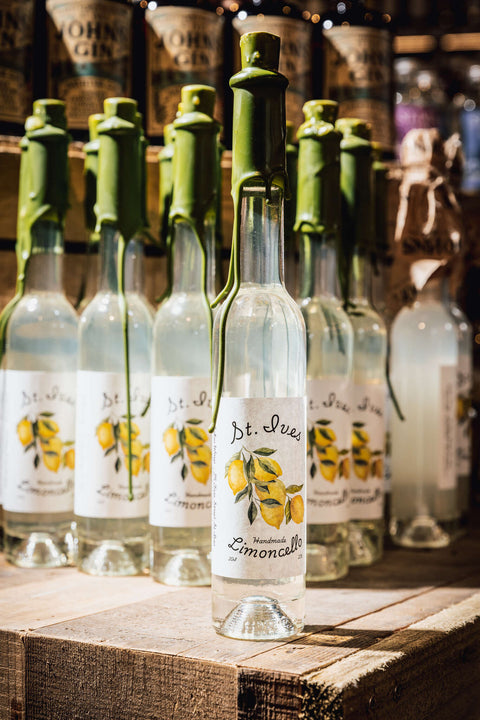 A row of clear glass bottles with white labels featuring yellow lemon illustrations and green stoppers, arranged on a rustic wooden surface with a warm, dimly lit background.