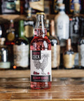 A bottle of Fruity Cock Berry Cider stands on a wooden bar counter with a blurred background of liquor bottles. The glass bottle contains pink liquid and has a white label with a black rooster illustration and red text.