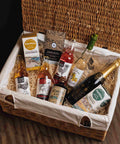 A wicker gift basket lined with a cream canvas bag and filled with assorted gourmet snacks and drinks, including a bottle of Johnnie Walker, a bottle of Pernic, a box of Furniss Shortbread, and a bottle of Scotch whisky, arranged on a wooden floor.