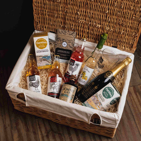 A wicker gift basket lined with a cream canvas bag and filled with assorted gourmet snacks and drinks, including a bottle of Johnnie Walker, a bottle of Pernic, a box of Furniss Shortbread, and a bottle of Scotch whisky, arranged on a wooden floor.
