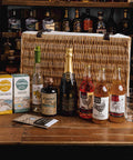 A wicker picnic-style gift hamper filled with assorted spirits and snacks, displayed on a wooden bar counter in front of shelves of liquor bottles. Visible items include a bottle of Johnnie Gin, a bottle of Duff Fine Gin, a bottle of Craft Cider, two Furals boxes, a box of Shortbread, and a glass.