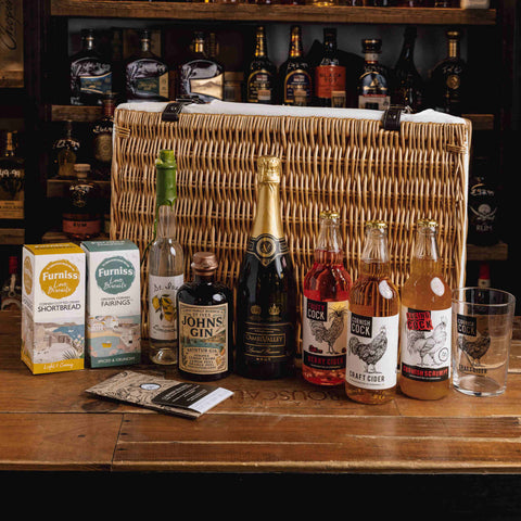A wicker picnic-style gift hamper filled with assorted spirits and snacks, displayed on a wooden bar counter in front of shelves of liquor bottles. Visible items include a bottle of Johnnie Gin, a bottle of Duff Fine Gin, a bottle of Craft Cider, two Furals boxes, a box of Shortbread, and a glass.
