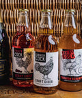 Three glass bottles of craft cider lined up on a wooden surface in front of a wicker basket. The bottles contain pink, amber, and golden-amber liquids, with white labels featuring black rooster illustrations and text including 'Fruity Cock', 'Cornish Cock', and 'Racing Cock'.