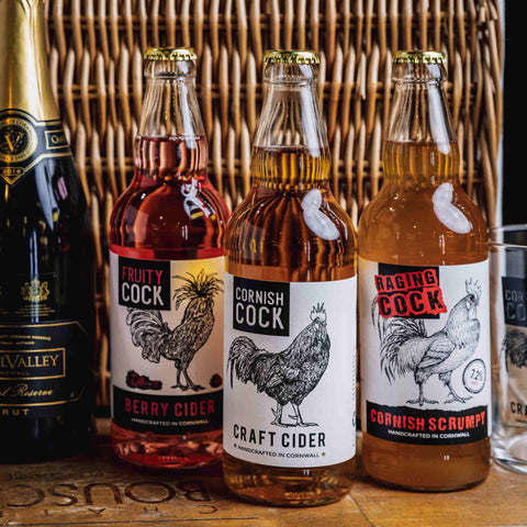 Three glass bottles of craft cider lined up on a wooden surface in front of a wicker basket. The bottles contain pink, amber, and golden-amber liquids, with white labels featuring black rooster illustrations and text including 'Fruity Cock', 'Cornish Cock', and 'Racing Cock'.