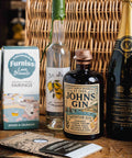 A styled product lineup on a wooden table featuring a bottle of St. Ives John's Gin, a bottle of Camell Valley Brut, Furniss Love Biscuits packaging, a lemon meringue sandwich cookie, and a branded card, set against a wicker basket background.