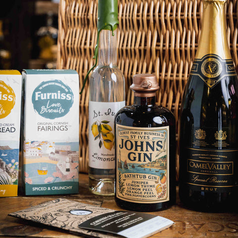 A styled product lineup on a wooden table featuring a bottle of St. Ives John's Gin, a bottle of Camell Valley Brut, Furniss Love Biscuits packaging, a lemon meringue sandwich cookie, and a branded card, set against a wicker basket background.
