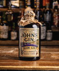 A bottle of John's Gin Sloe Gin sits on a wooden bar counter with a blurred bar background of other bottles. The bottle is dark glass, sealed with a rustic brown paper band, and features a vintage-style cream label with black text and a purple and orange graphic.