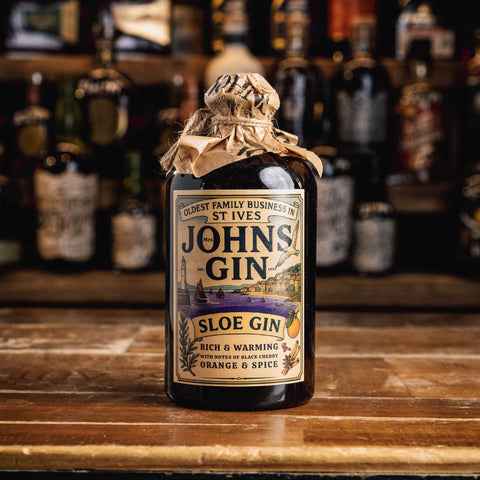 A bottle of John's Gin Sloe Gin sits on a wooden bar counter with a blurred bar background of other bottles. The bottle is dark glass, sealed with a rustic brown paper band, and features a vintage-style cream label with black text and a purple and orange graphic.