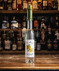 A bottle of St. Louis Lemonade stands on a wooden bar counter with a blurred background of shelves filled with liquor bottles.