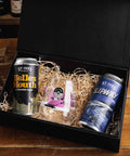 A black gift box filled with shredded paper packaging on a wooden table, containing two St. Ives Brewing cans and a small clear glass with a pink and white label.
