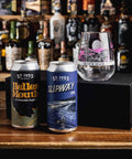 Two St. Ives Brewing cans, Helles Mouth and Slipway IPA, displayed on a wooden bar counter next to a custom engraved clear glass, with a black box and a blurred background of liquor bottles.