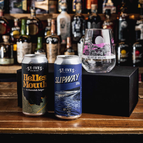 Two St. Ives Brewing cans, Helles Mouth and Slipway IPA, displayed on a wooden bar counter next to a custom engraved clear glass, with a black box and a blurred background of liquor bottles.