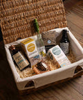 A wicker picnic basket with a cream canvas liner filled with gourmet food items, including bottles of Johnnie Walker and Skyyra, Furnace Shopbread, and a chocolate bar, arranged on a wooden floor.
