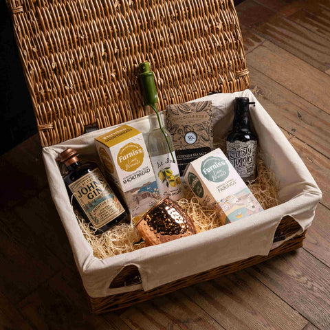 A wicker picnic basket with a cream canvas liner filled with gourmet food items, including bottles of Johnnie Walker and Skyyra, Furnace Shopbread, and a chocolate bar, arranged on a wooden floor.