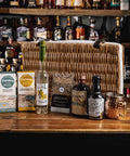 A curated selection of gourmet snacks and spirits arranged on a wooden bar counter in front of a large woven basket, with shelves of liquor bottles in the background. Visible products include Furnise Creamery Farrowgs and Sweetbreads, Mr.