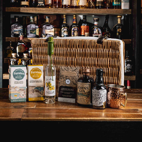 A curated selection of gourmet snacks and spirits arranged on a wooden bar counter in front of a large woven basket, with shelves of liquor bottles in the background. Visible products include Furnise Creamery Farrowgs and Sweetbreads, Mr.