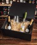 A black gift box filled with shredded paper and three bottles of sparkling wine or champagne, plus a clear wine glass, displayed on a wooden table with shelves of liquor bottles in the background.
