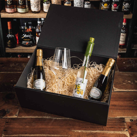 A black gift box filled with shredded paper and three bottles of sparkling wine or champagne, plus a clear wine glass, displayed on a wooden table with shelves of liquor bottles in the background.