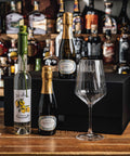 A gift set of three bottles of J'1s Bar ROCCO wine and sparkling wine with a clear wine glass, arranged on a wooden table in front of a black box and a blurred background of liquor bottles.