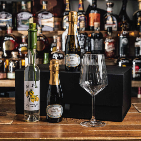 A gift set of three bottles of J'1s Bar ROCCO wine and sparkling wine with a clear wine glass, arranged on a wooden table in front of a black box and a blurred background of liquor bottles.