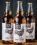 Three glass bottles of Cornish Cock Craft Cider arranged in a row on a wooden surface against a dark background. The center bottle is in sharp focus, with the left and right bottles softly blurred.