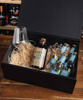 A black gift box filled with crinkle paper packaging on a wooden table, containing a bottle of John Gin, several bottles of Lava sparkling wine, and a clear wine glass.