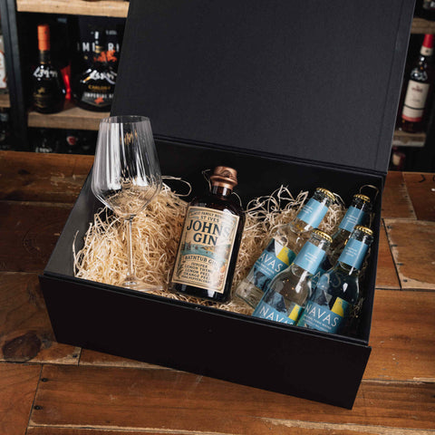 A black gift box filled with crinkle paper packaging on a wooden table, containing a bottle of John Gin, several bottles of Lava sparkling wine, and a clear wine glass.